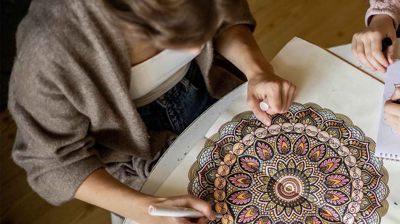 Een vrouw zit aan tafel en tekent een mandala. 